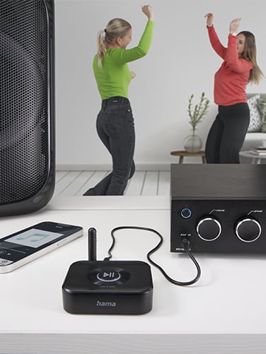 Hama Bluetooth® adapter "Link.it solo", a hi-fi system and a smartphone can be seen on a chest of drawers. The smartphone is connected to the adapter via Bluetooth and plays music on the hi-fi system. Two young ladies are dancing in the background.